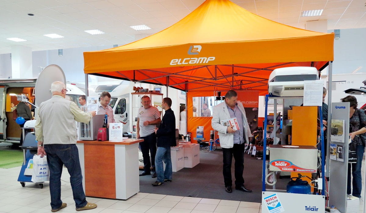 Faltzelt 3x3m Messestand Faltzelt 3x3m Messestand von elcamp mit orangefarbenem Dach, Besucher am Stand, Produkte ausgestellt.