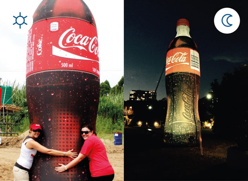 Blow Up Easy Inflate Ad Coca Cola Zwei Frauen umarmen eine große Coca-Cola-Flasche, die bei Tag und Nacht beleuchtet ist.