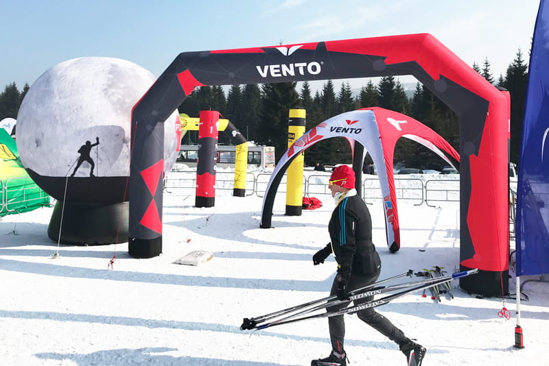 Skifahrer unter VENTO aufblasbarem Tor Ein Skifahrer mit Skistöcken geht unter einem aufblasbaren Tor mit dem Logo 'VENTO' hindurch, umgeben von Schnee und weiteren aufblasbaren Elementen.