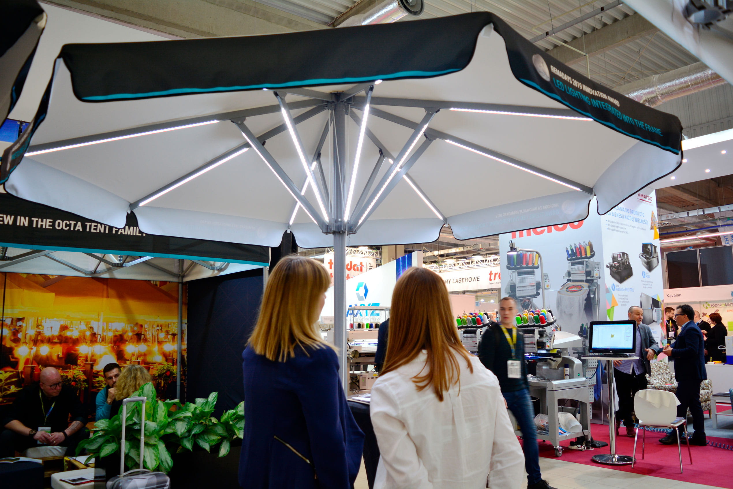 Sonnenschirm Expert Led Messe Zwei Personen betrachten den Sonnenschirm Expert Led Messe am Stand, umgeben von Ausstellungsständen.