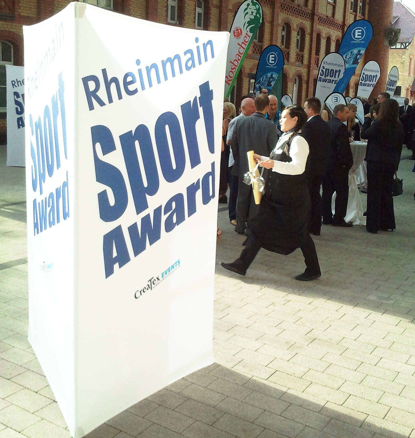 Werbesäule Clickboard Vertical Sport Award Werbesäule mit der Aufschrift 'Rheinmain Sport Award' auf einer Veranstaltung, umgeben von Gästen in formeller Kleidung.