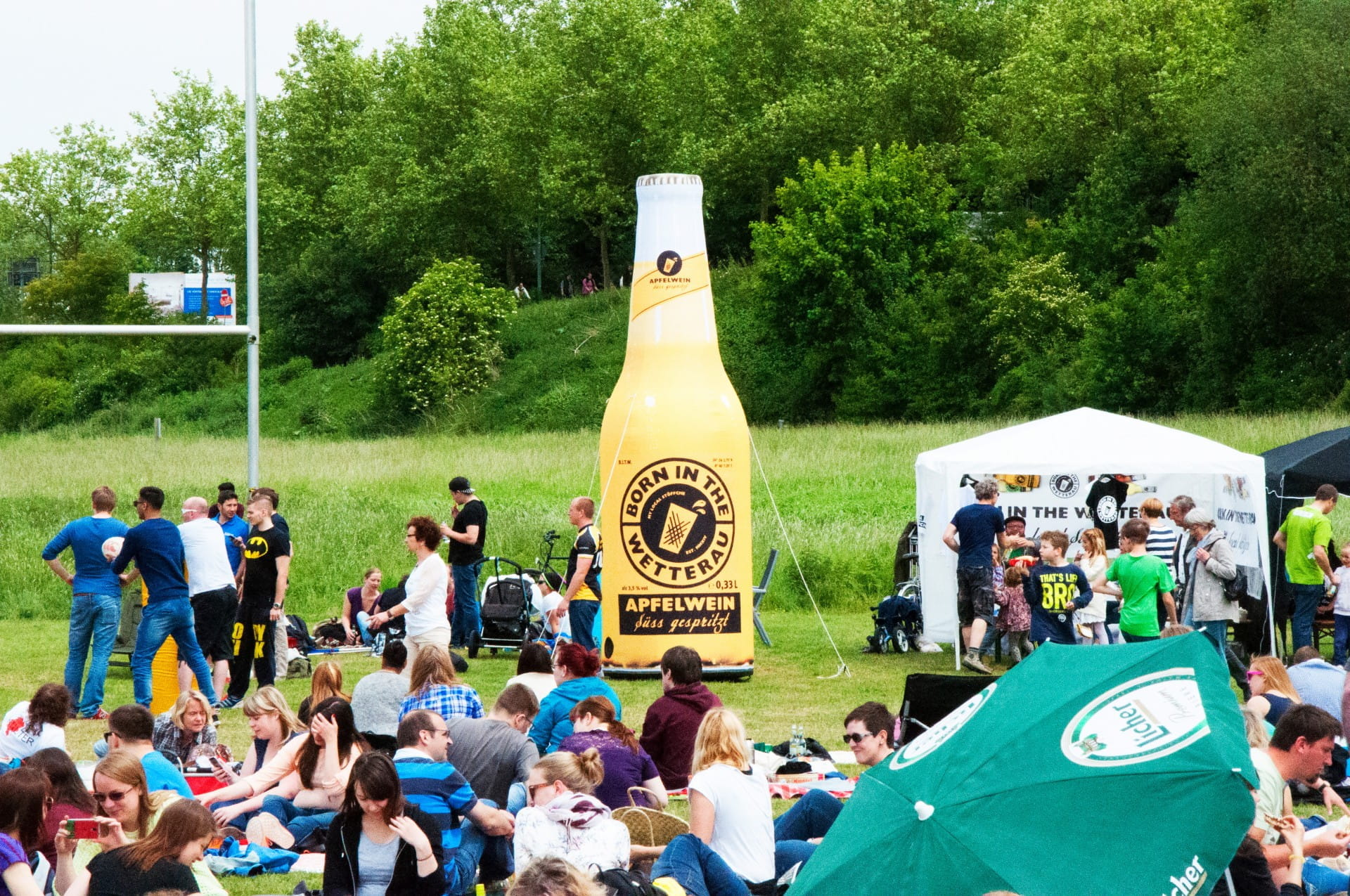 Blow Up Easy Inflate Ad Flasche Born In The Wetterau Auf einem Festivalgelände steht eine große aufblasbare Flasche mit der Aufschrift 'Born In The Wetterau'. Menschen sitzen auf dem Gras und genießen die Veranstaltung.