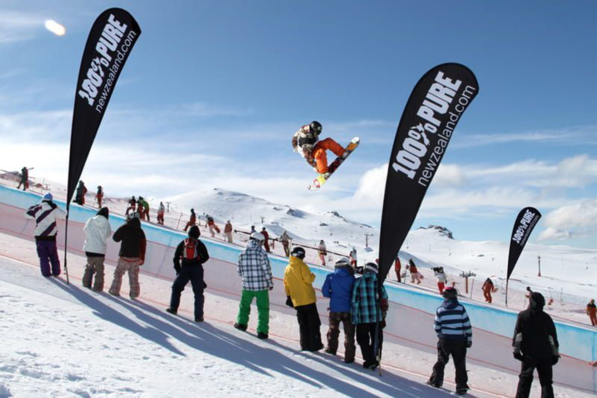 Snowboardfahrer macht einen großen Sprung vor Publikum auf einer Piste, mit drei großen Beachflags
