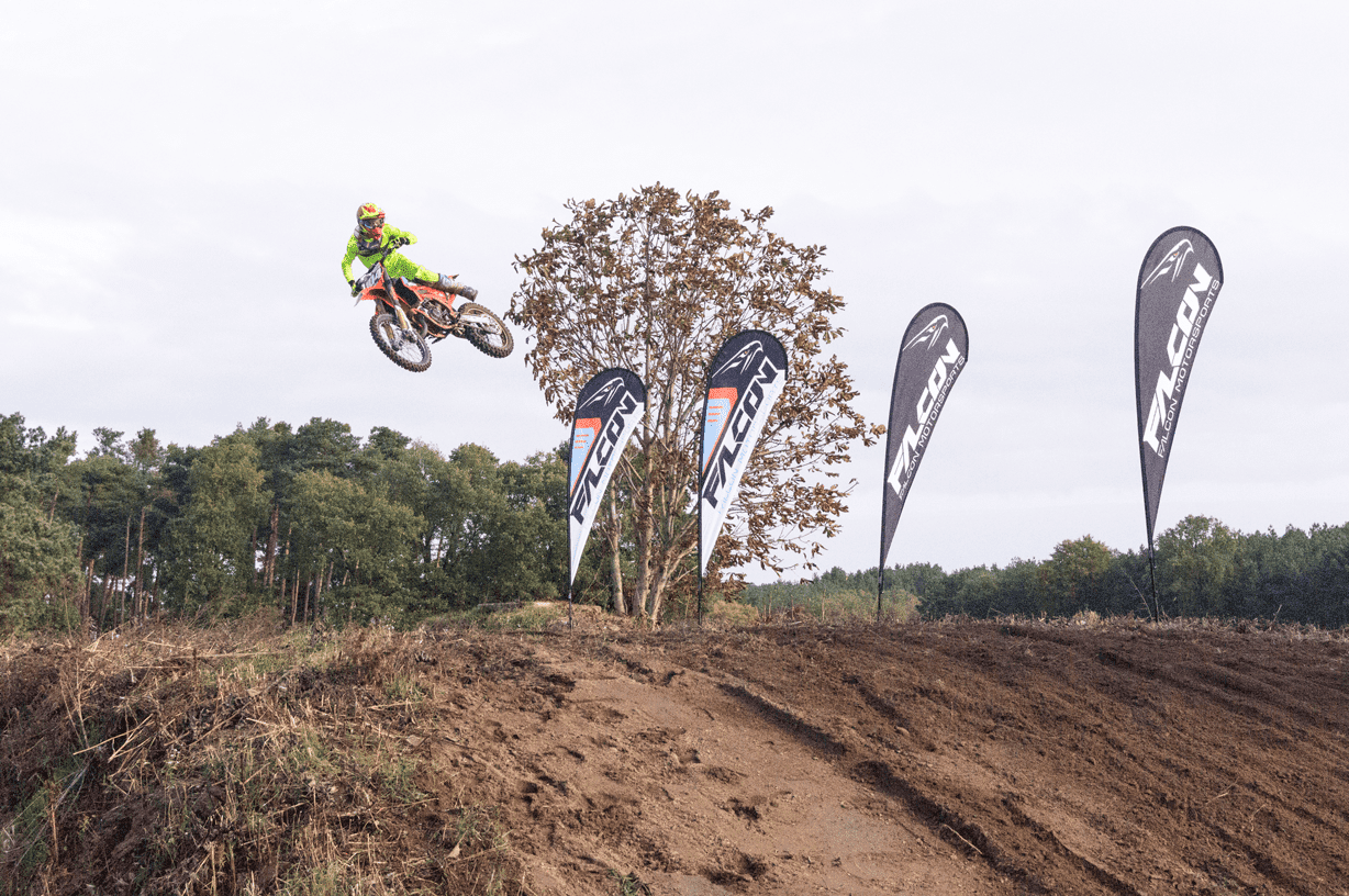 Falcon Motorsports Jump Beachflag Easyflag Drop Motorradfahrer springt mit einem Motorrad über einen Hügel, umgeben von Falcon Beachflags.