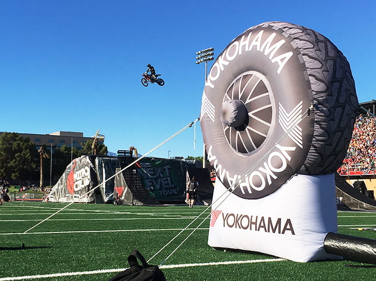 Aufblasbare Yokohama-Werbung bei einem Sportevent Aufblasbare Werbung in Form eines Yokohama-Reifens auf einem Sportfeld, während ein Motorradfahrer in der Luft springt.