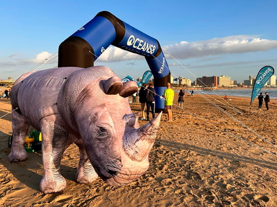 Aufblasbares Nashorn am Strand Ein großer, aufblasbarer Nashorn steht am Strand unter einem Bogen mit der Aufschrift 'OCEANSB'. Im Hintergrund sind Menschen und Gebäude sichtbar.