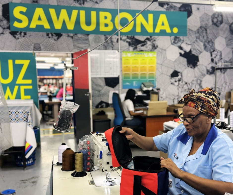 Frau bei der Arbeit in einer Näherei mit 'SAWUBONA.' im Hintergrund Eine Frau in blauer Arbeitskleidung näht an einer Maschine in einer Fabrik. Im Hintergrund steht 'SAWUBONA.' an der Wand.