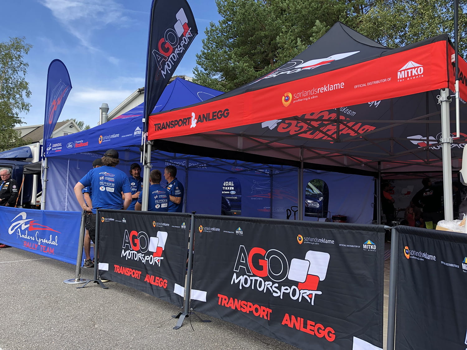 Rennsportbande Motorsport Zelt und Banner von AGO Motorsport bei einer Motorsportveranstaltung, mit Teammitgliedern in blauen Shirts.