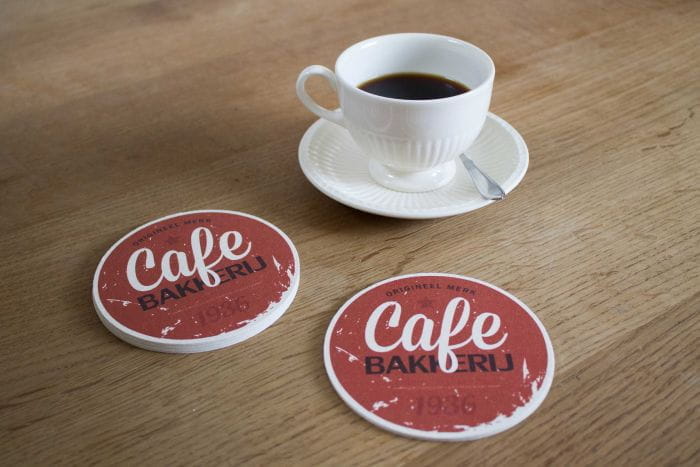 Untersetzer Gastronomie Zwei runde Untersetzer mit der Aufschrift 'Cafe Bakudrij 1976' neben einer Tasse Kaffee auf einem Holztisch.