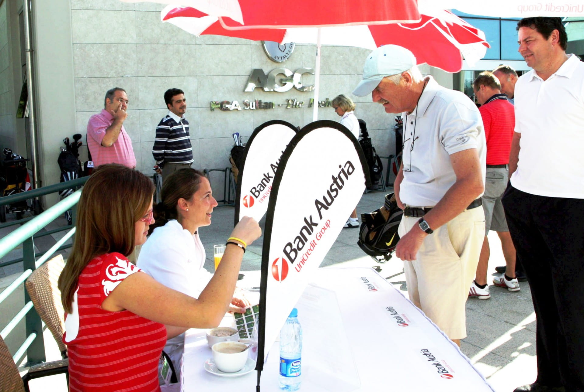 Tischaufstelle von Bank Austria bei einem Event Zwei Frauen am Tisch mit Bank Austria-Logo beraten einen Mann, während andere Personen im Hintergrund stehen.