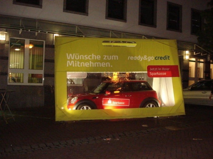 Autotasche Bei Nacht Eine Autotasche mit einem Mini Cooper darin, beleuchtet bei Nacht. Auf der Tasche steht 'Wünsche zum Mitnehmen. ready&go credit. Jetzt in Ihrer Sparkasse'.