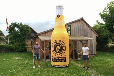 Aufblasbare Flasche 'Born In The Wetterau' Zwei Männer halten eine große aufblasbare Flasche mit dem Aufdruck 'Born In The Wetterau Apfelwein' vor einem Holzgebäude.