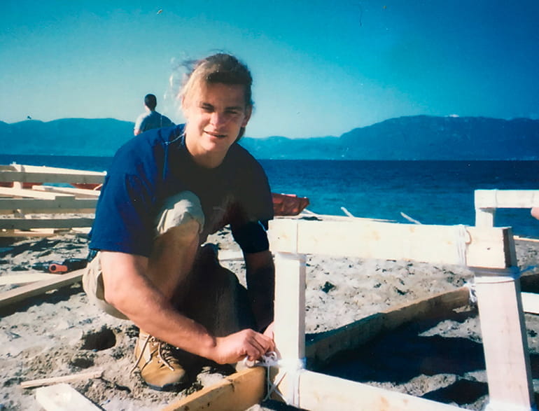 Christian Decker beim Incentiv Bootsbau am Strand Ein junger Mann mit langen Haaren kniet am Strand und arbeitet an einem Holzrahmen für den Bootsbau.