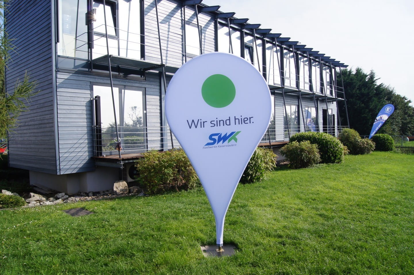 Sonderprojekte Standortmarker Standortmarker mit der Aufschrift 'Wir sind hier.' und dem Logo von SWK, auf einer Wiese vor einem modernen Gebäude.
