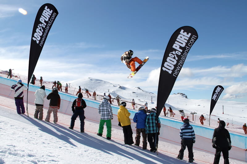 Snowboard-Event mit Beachflags Snowboarder springt über eine Halfpipe, umgeben von Zuschauern und Beachflags mit dem Text '100% PURE'.