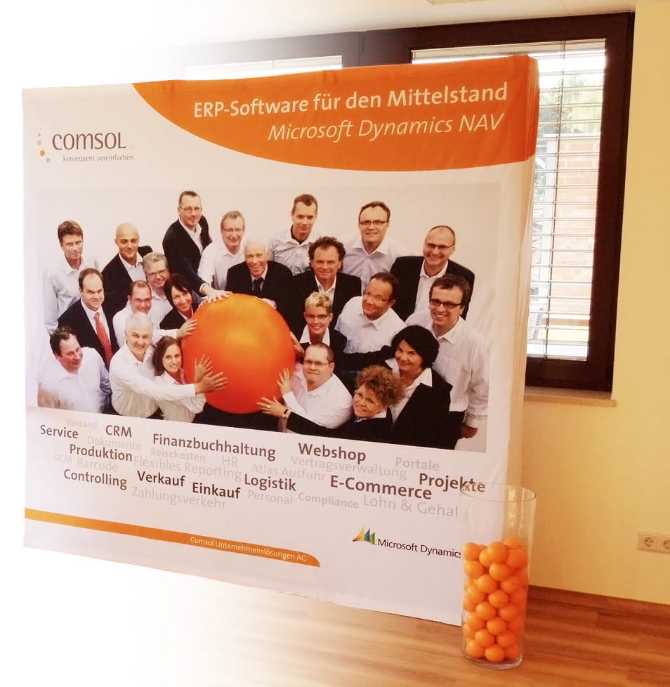 Messewand Quickup Stoffwand Comsol Messewand mit dem Produktnamen 'Quickup Stoffwand Comsol', zeigt ein Team von Personen vor einem großen orangefarbenen Ball, mit Text über ERP-Software für den Mittelstand und Microsoft Dynamics NAV.