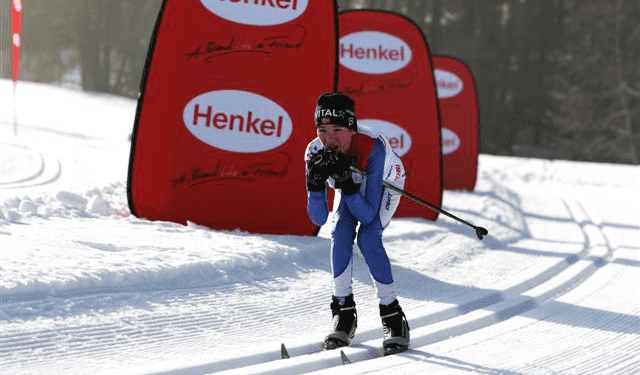 Werbesäule Quicktower Henkel Skilangläufer in blauer und weißer Sportkleidung, der auf einer schneebedeckten Strecke fährt, mit Werbesäulen von Henkel im Hintergrund.
