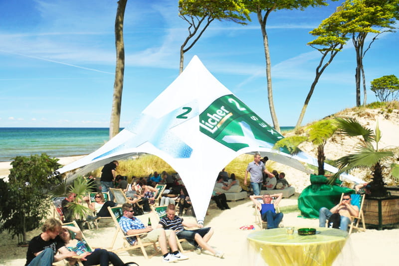 Sternzelt Beach am Strand Sternzelt Beach von ENTDECKER, ein großes, weißes Zelt mit grünen und blauen Akzenten, umgeben von Menschen am Strand.