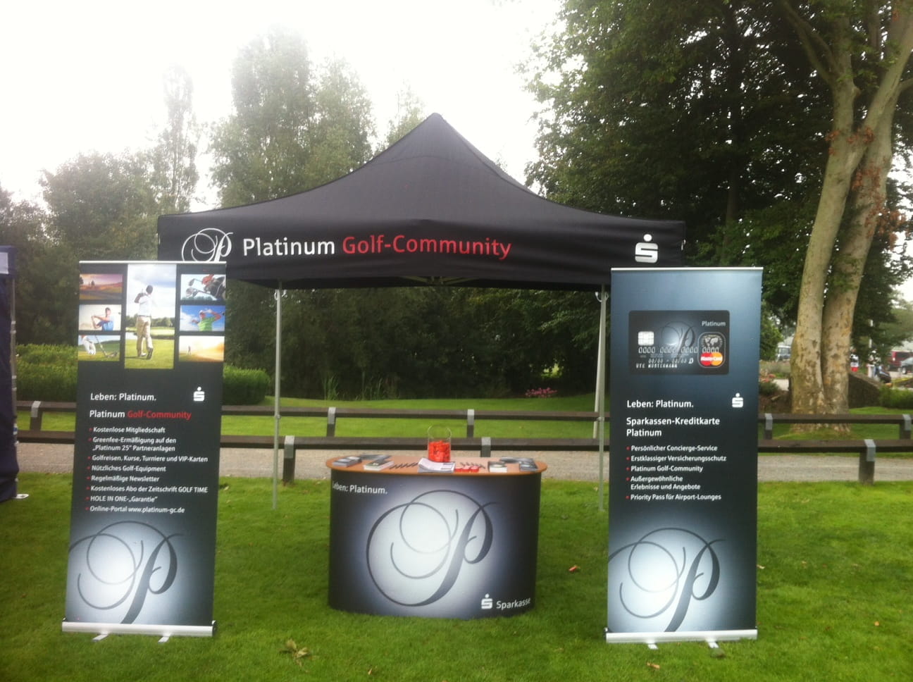Golf Outdoor Stand Stand der Platinum Golf-Community mit zwei Roll-Ups und einem Tisch im Freien, umgeben von Bäumen.