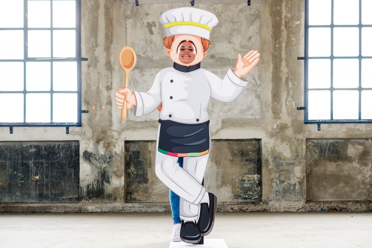 Inhaker Aufsteller Koch Fotofigur Aufsteller einer Kochfigur mit Kochmütze und Kochlöffel, steht in einem Raum mit großen Fenstern.