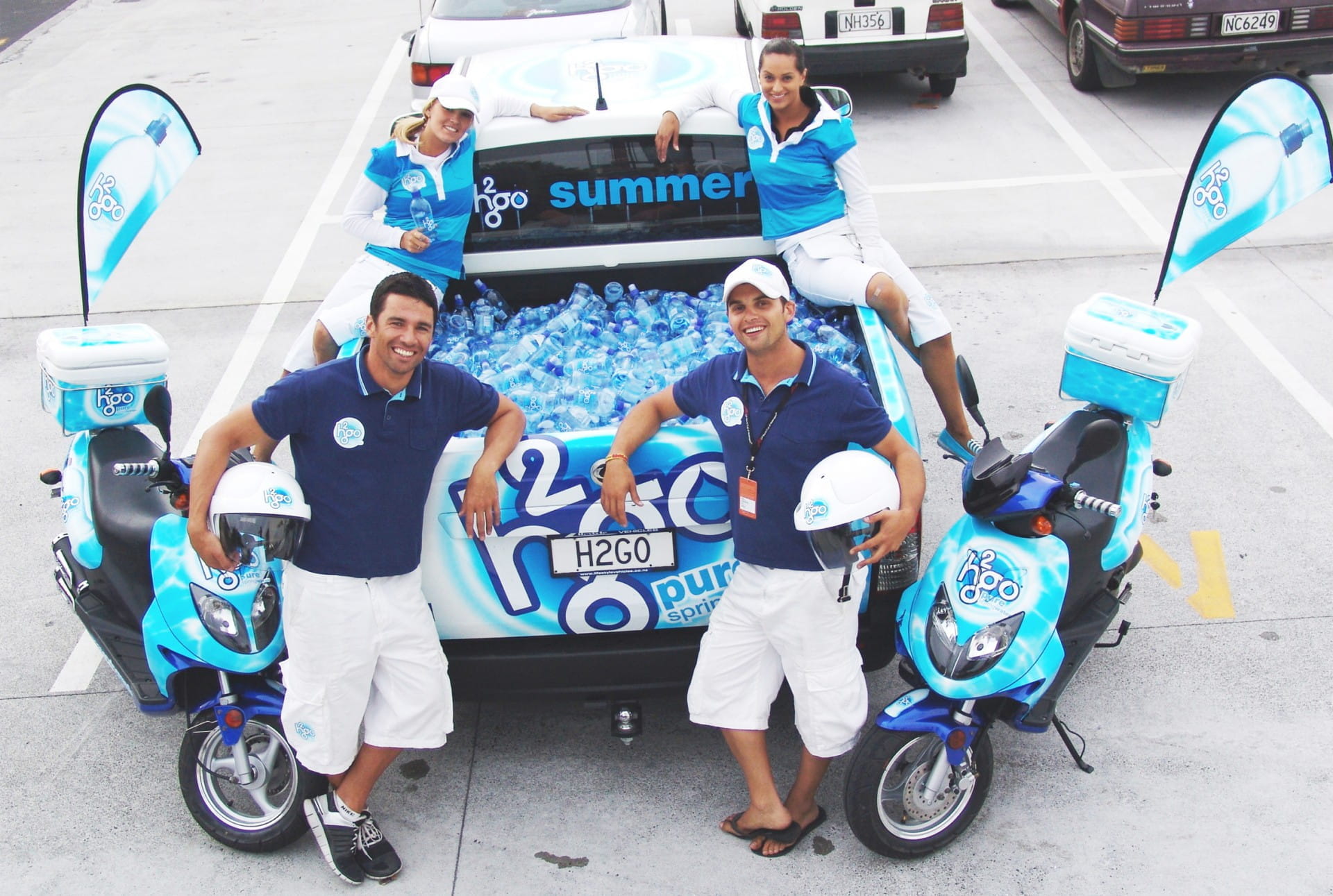 H2GO Promotionsteam vor Fahrzeug Drei Personen in blauen H2GO-T-Shirts stehen vor einem Fahrzeug mit Wasserflaschen und H2GO-Logo. Zwei Motorräder sind sichtbar.