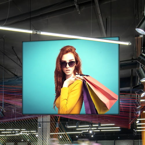 Deckenhänger Easyframe im Modegeschäft Deckenhänger Easyframe im Modegeschäft mit einer Frau in gelbem Oberteil und Sonnenbrille, die bunte Einkaufstaschen trägt.