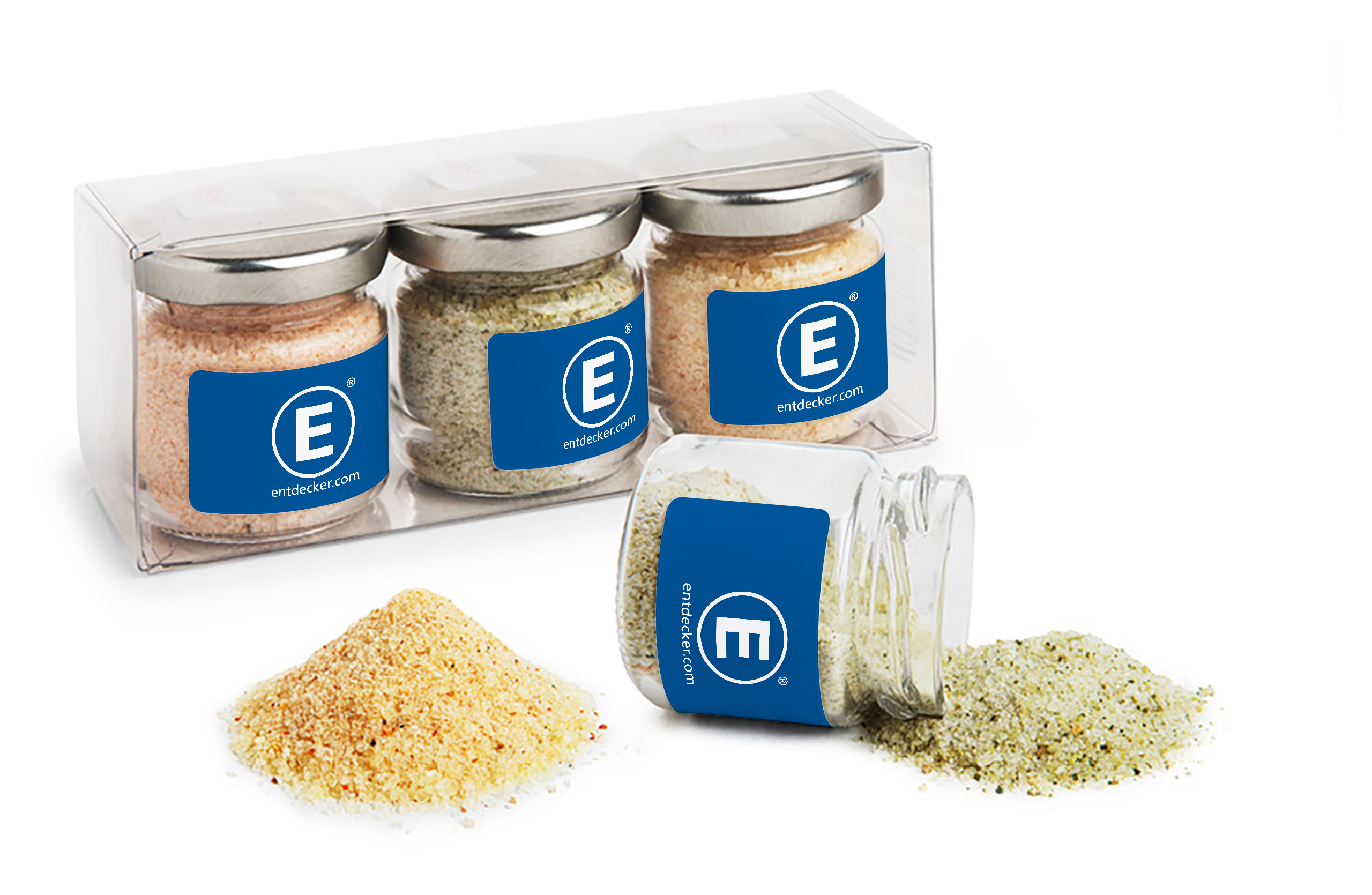 Drei kleine Salzgläser mit unterschiedlichen den Gourmetsalz Sorten Chili-Ingwer, Dill-Petersilie und Masala-Curry in einer transparenten Box, daneben ein umgestürztes Glas mit ausgestreutem Salz.