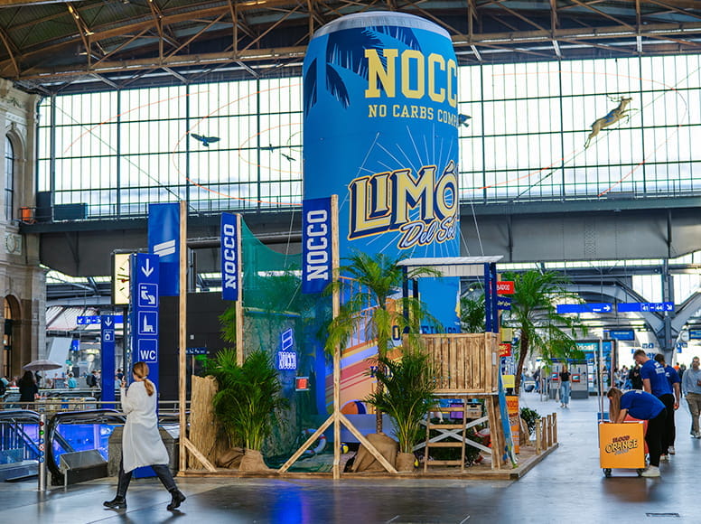Aufblasbare Werbung Limonade Slider von NOCCO Aufblasbare Werbung für NOCCO Limonade in einem Bahnhof, umgeben von Pflanzen und Holzdekoration.
