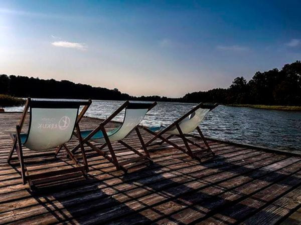 Liegestuhl See 600x600 Drei Liegestühle aus Holz mit transparenten Stoffen auf einem Holzsteg am Wasser, umgeben von Bäumen.