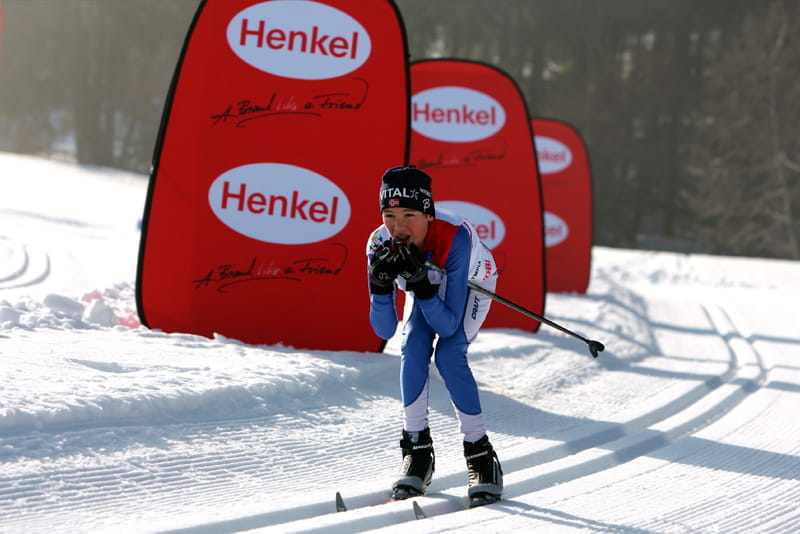 Skilanglauf-Wettbewerb mit Werbebanden von Henkel Skilangläufer in blauer Kleidung mit Skiern, der auf einer Loipe fährt, vor roten Werbebanden mit dem Logo von Henkel.