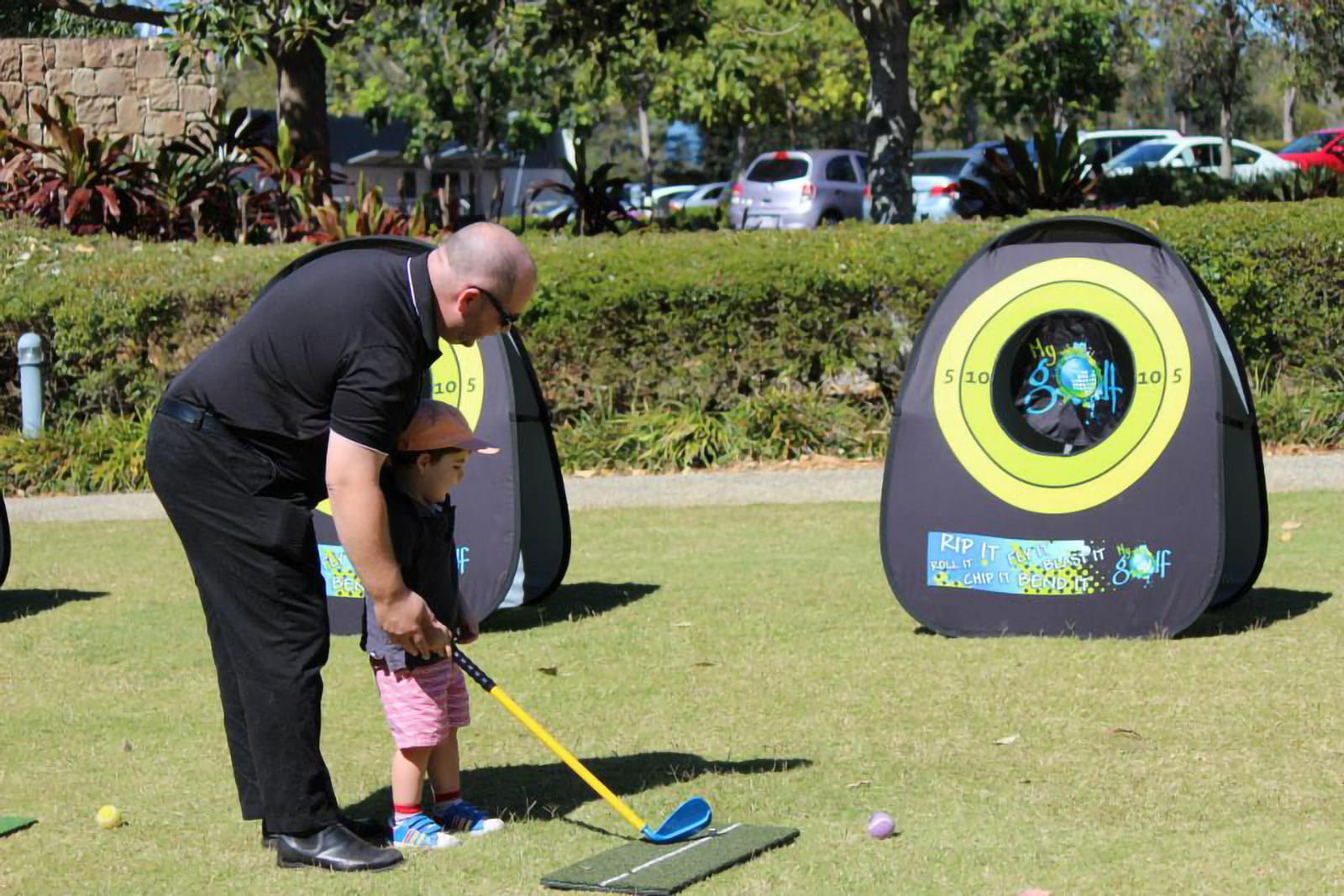 Zielscheibe Mobile Werbebande Quickboard Ein Erwachsener und ein Kind stehen vor einer Zielscheibe der mobilen Werbebande Quickboard im Freien. Der Erwachsene hält einen Golfschläger, während das Kind bereit ist, zu spielen.