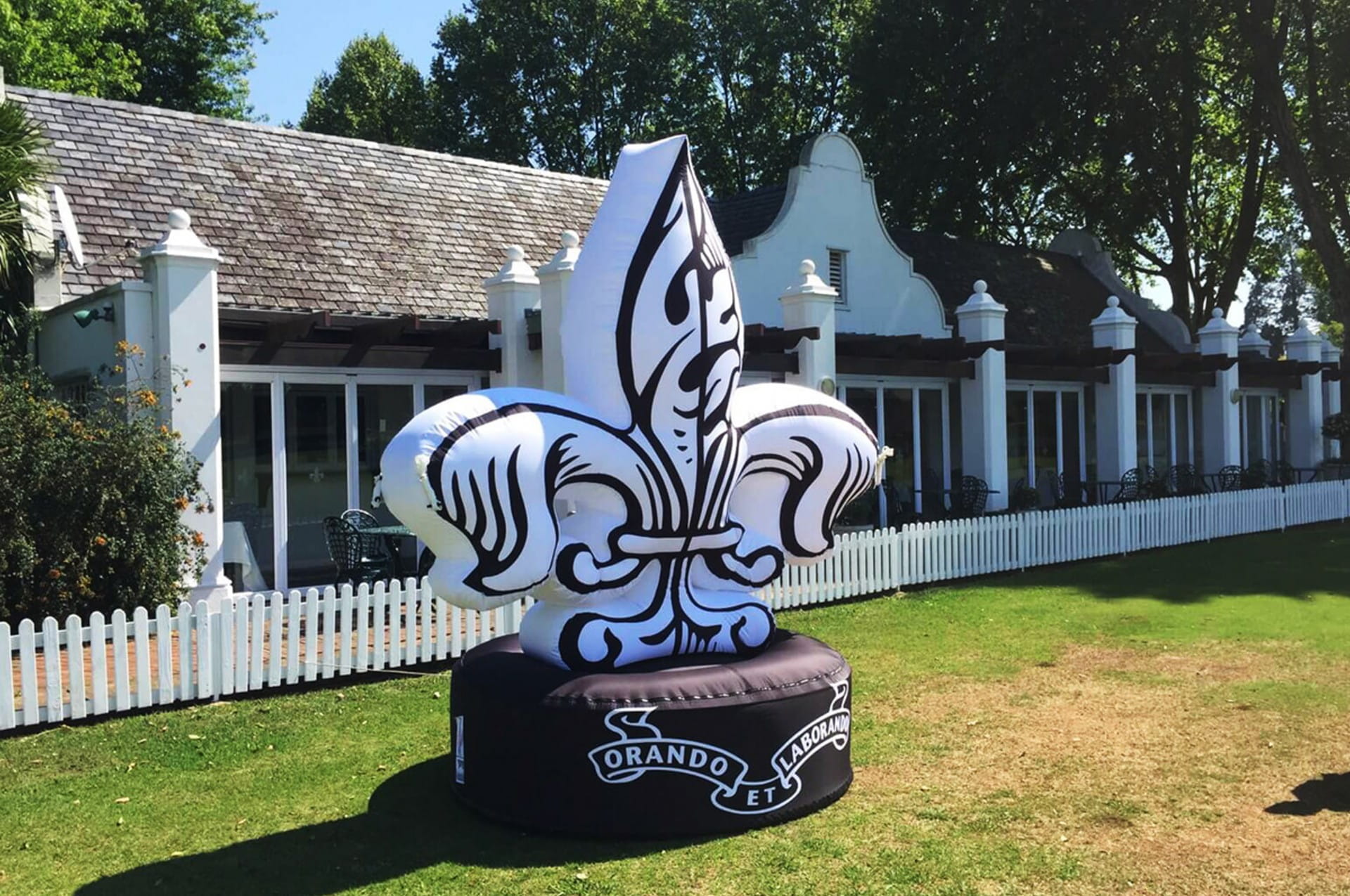 Blow Up Easy Inflate Ad Orando Aufblasbare Werbung in Form einer Lilie mit dem Schriftzug 'ORANDO ET' auf einem schwarzen Sockel, im Freien vor einem Gebäude.