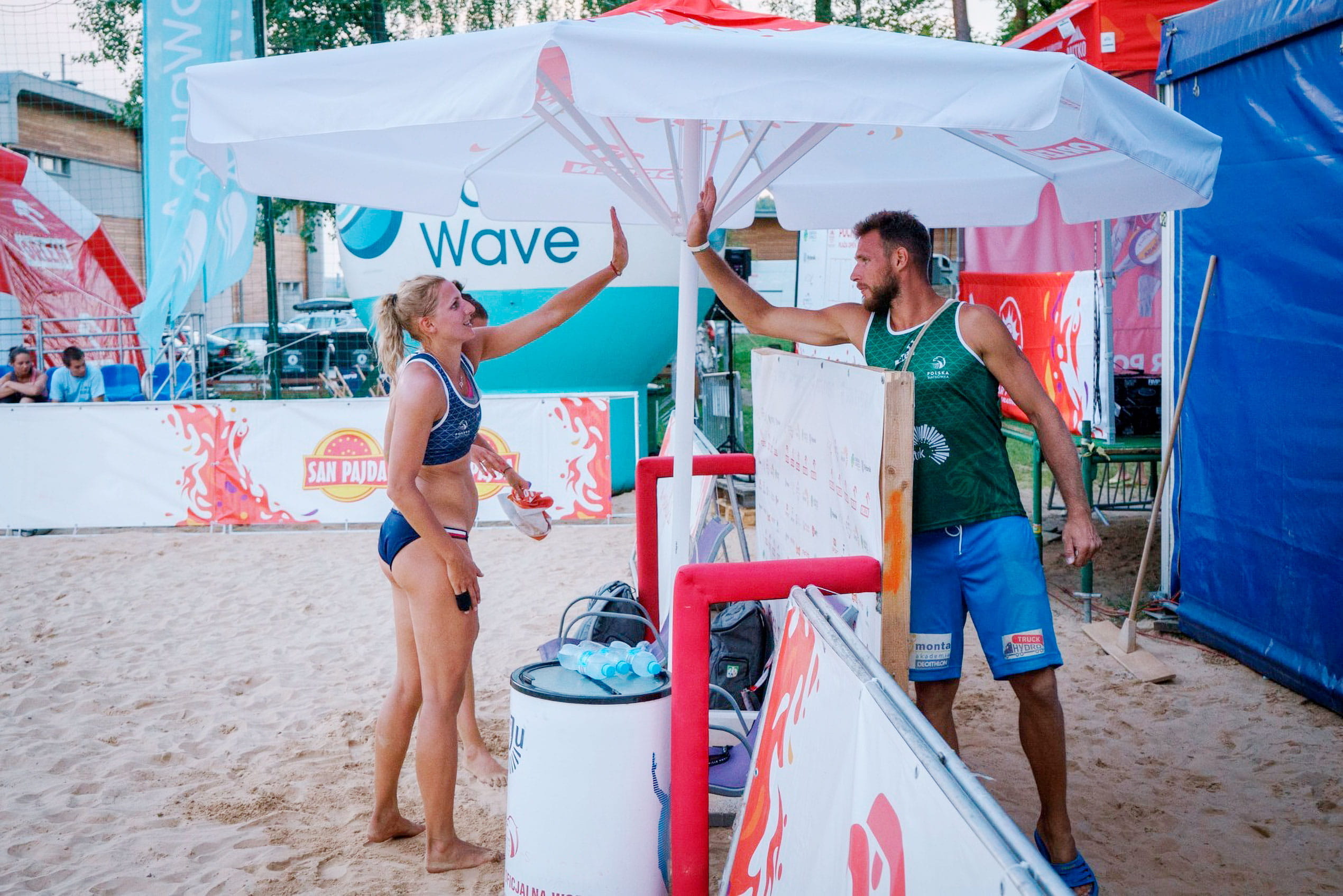 Sonnenschirm Expert Sport Event Zwei Personen unter einem Sonnenschirm Expert Sport Event am Strand, eine Frau in Sportkleidung und ein Mann in einem Tanktop, beide lächeln.