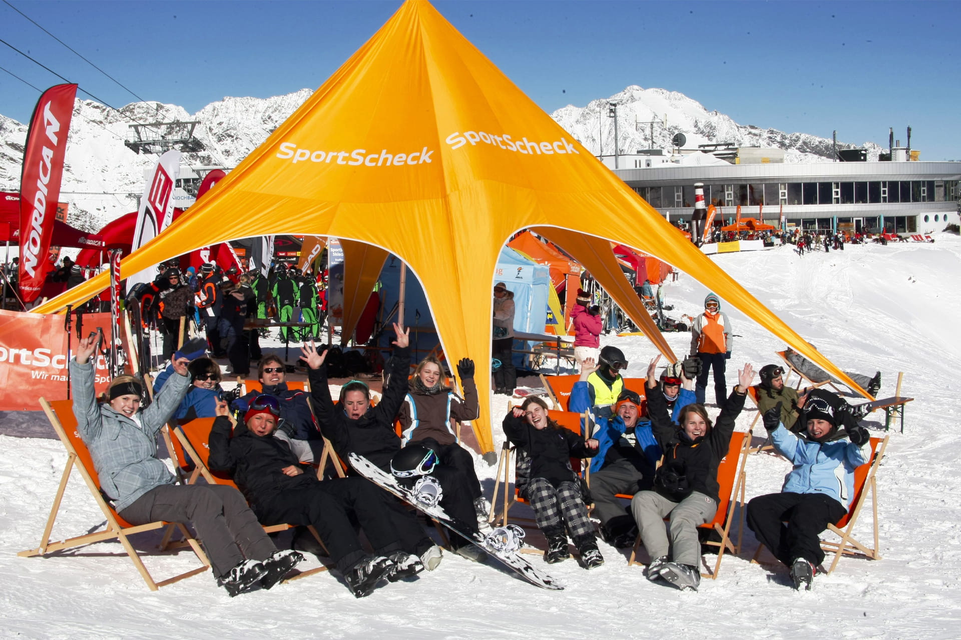 Orangenes sternenförmiges Zelt in einem Skigebiet mit vielen Menschen in Schneekleidung auf Liegestühlen daruntersitzend, Fahnen und Gebäude sowie ein Lift im Hintergrund