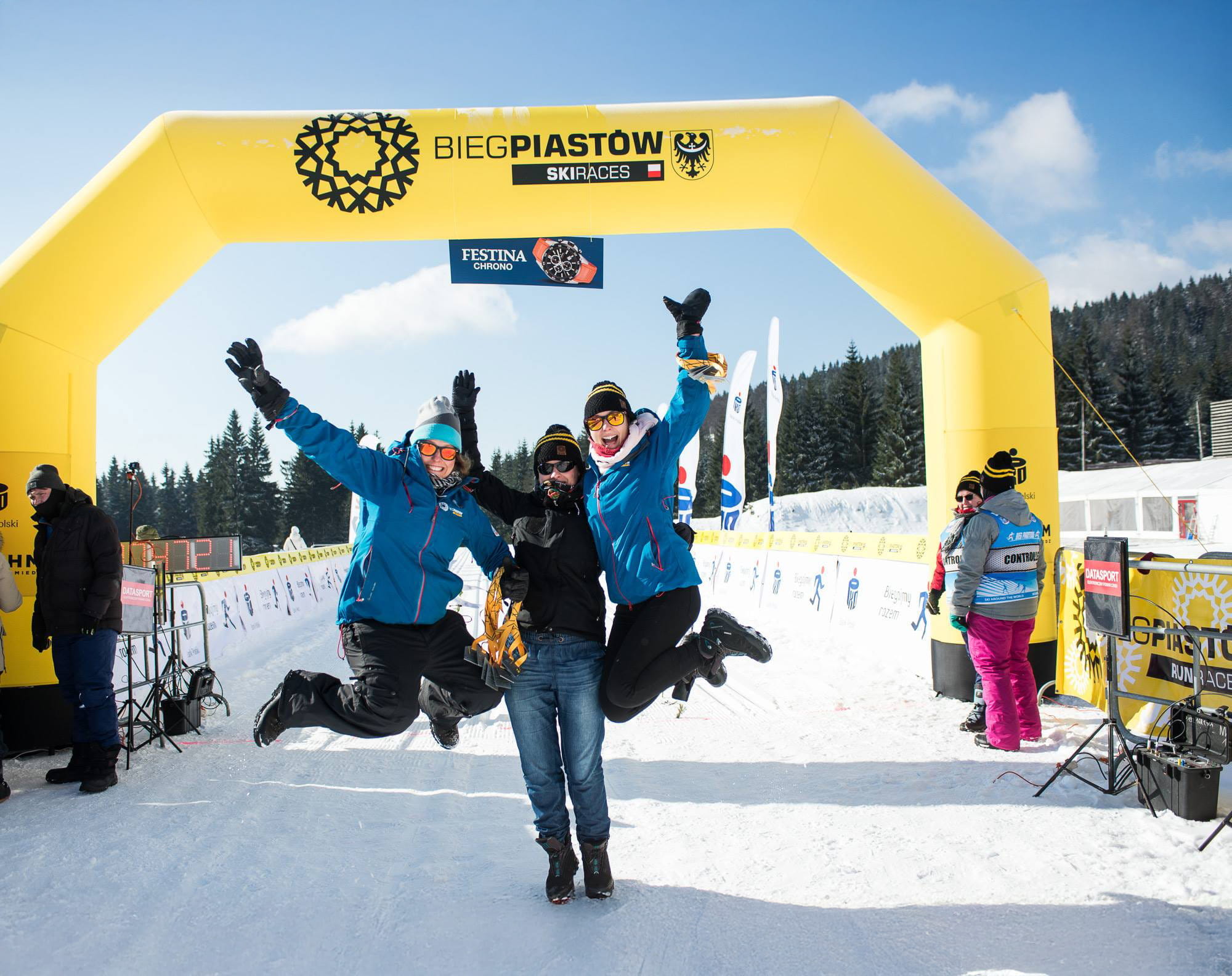 Vento Torbogen Wintersport Drei Personen feiern unter einem gelben Torbogen mit der Aufschrift 'BIEGPIASTOW SKIRACES' im Schnee.