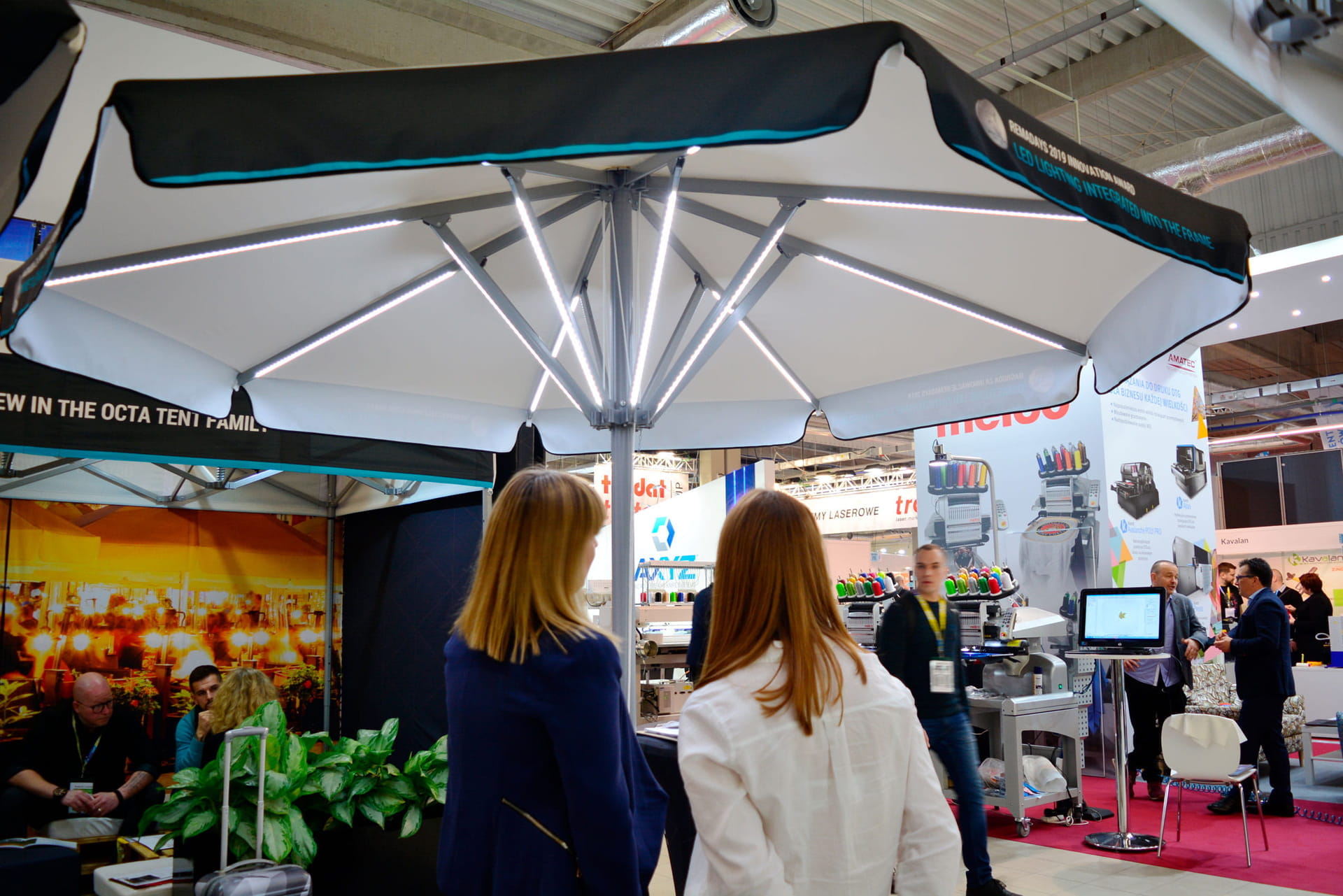 Sonnenschirm Expert Led Messe Zwei Personen betrachten den Sonnenschirm Expert Led Messe am Stand, umgeben von Ausstellungsständen.