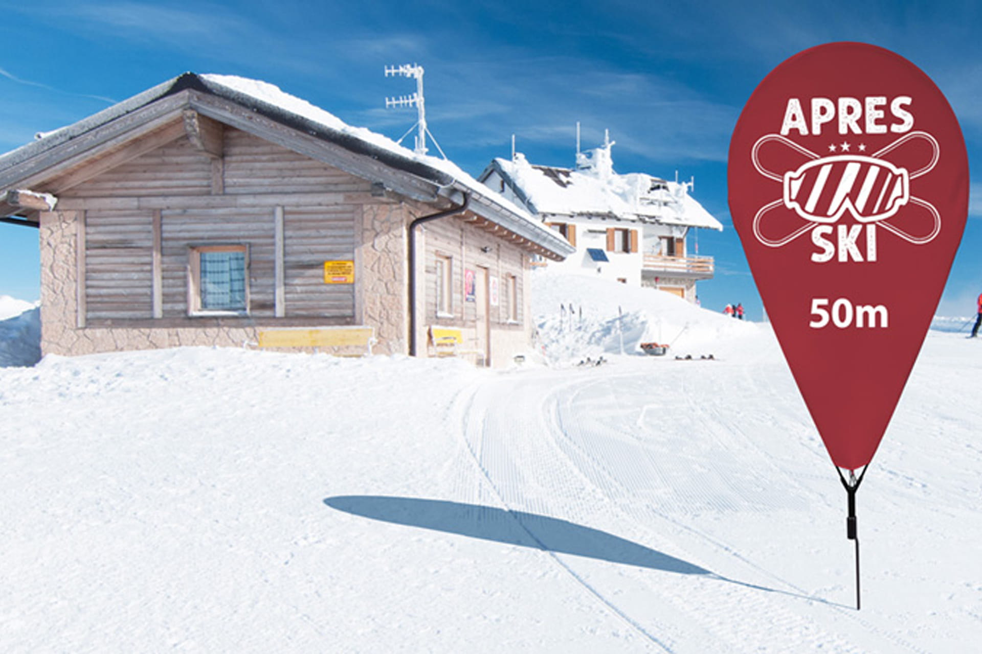 Eine dunkelrote Pinflag mit der Aufschrift "Apres Ski 50m" auf einer schneebedeckten Fläche vor zwei Skihütten