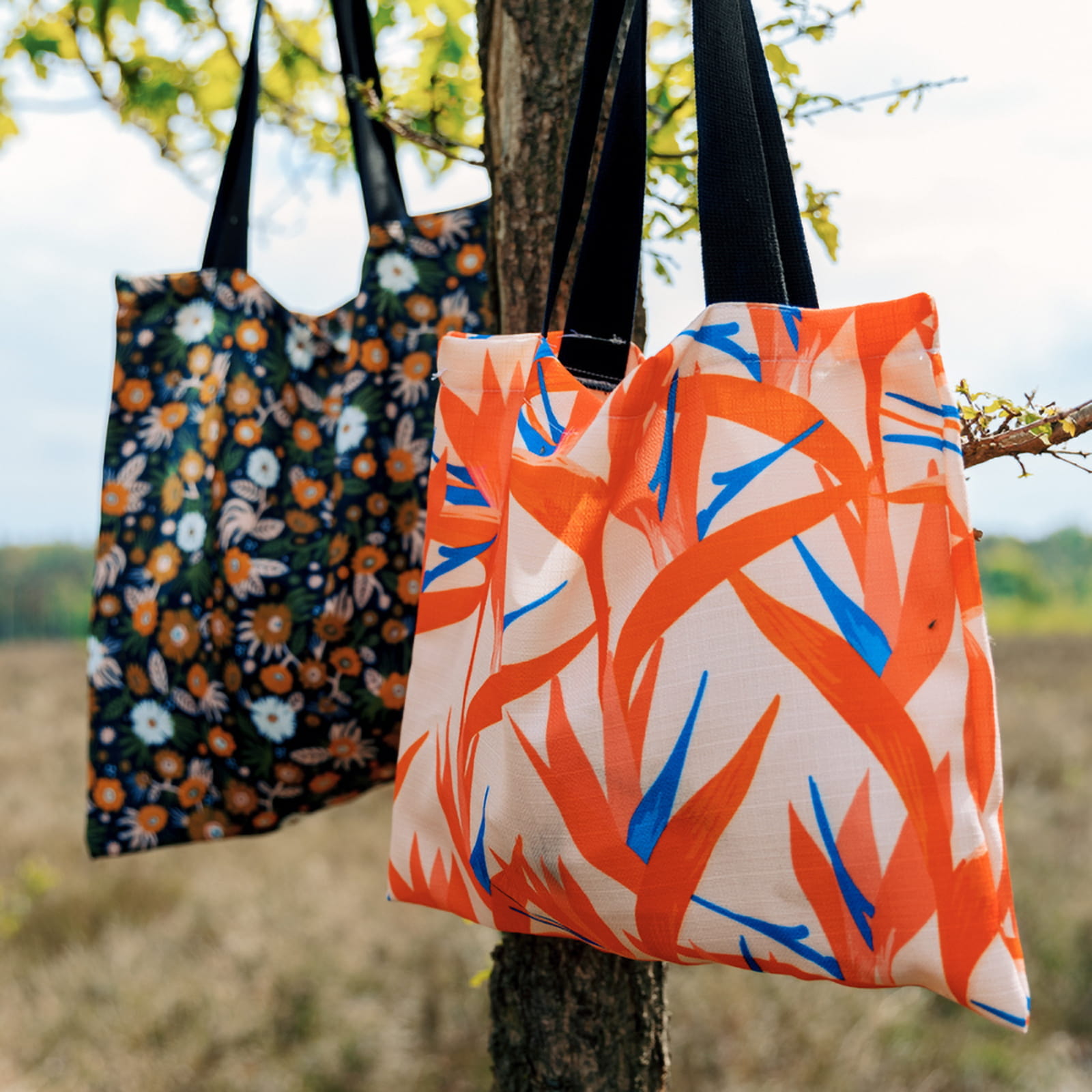 Tragetaschen bedruckt mit bunten Designs Zwei Tragetaschen von ENTDECKER mit bunten Designs, eine mit floralen Mustern und die andere mit abstrakten, orange-blauen Pflanzenmotiven, hängen an einem Baum.