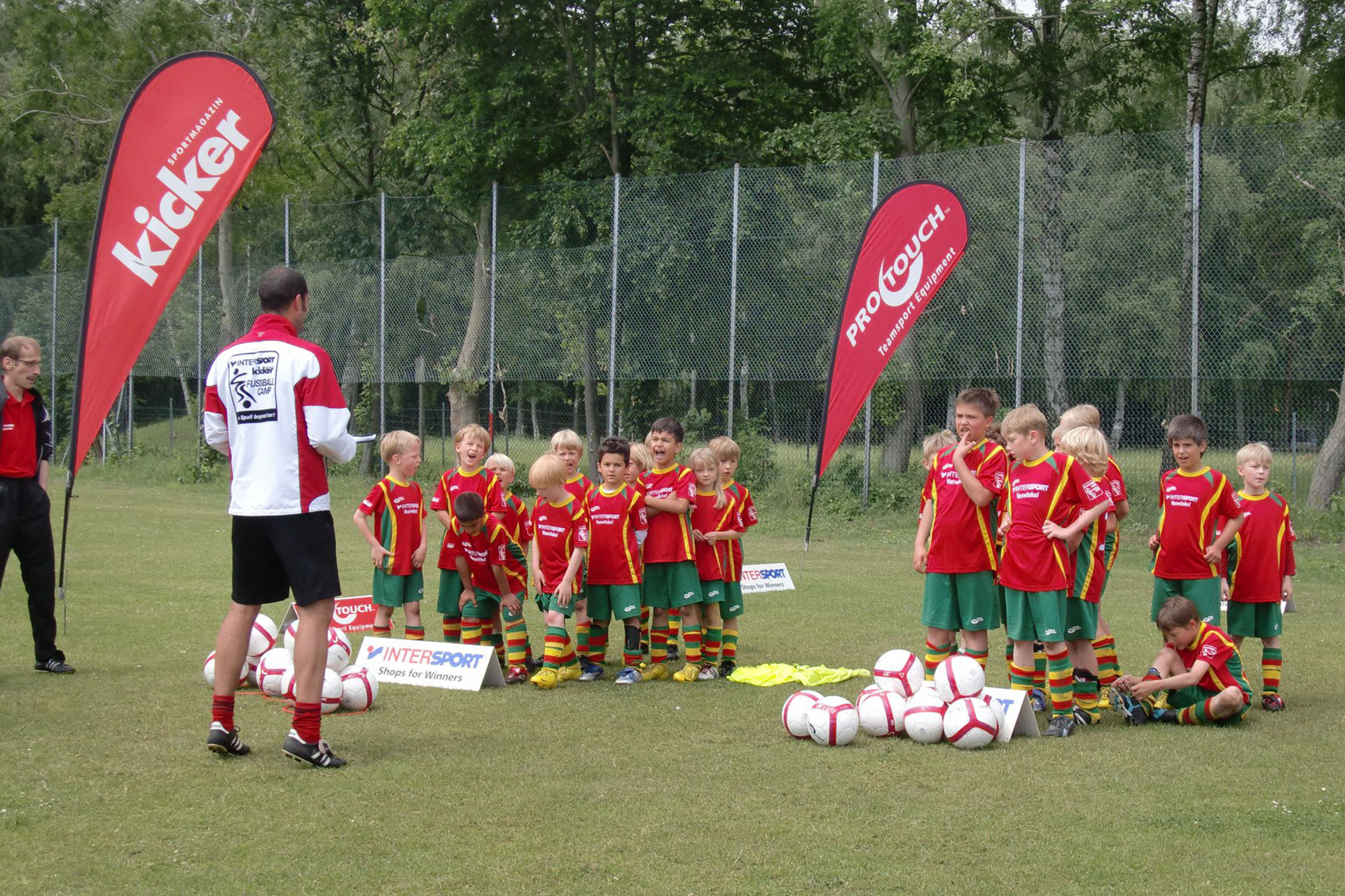 Kinder in Fußballausrüstung stehen vor dem Trainern mit einem Haufen Fußbälle und zwei Beachflags im Hintergrund