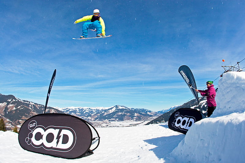 Snowboarder über Quickboard Winter Snowboarder springt über eine Werbebande "Quickboard Winter" von ENTDECKER, während eine Zuschauerin zuschaut. Winterlandschaft im Hintergrund.