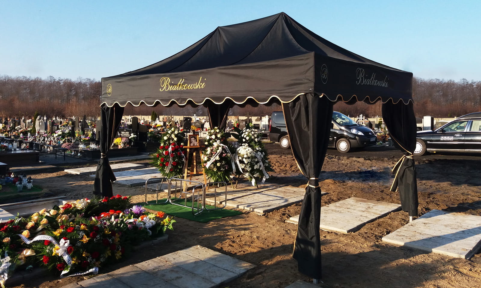 Mt Premium Beerdigungszelt Schwarzes Beerdigungszelt mit goldener Schrift, umgeben von Blumenarrangements auf einem Friedhof.