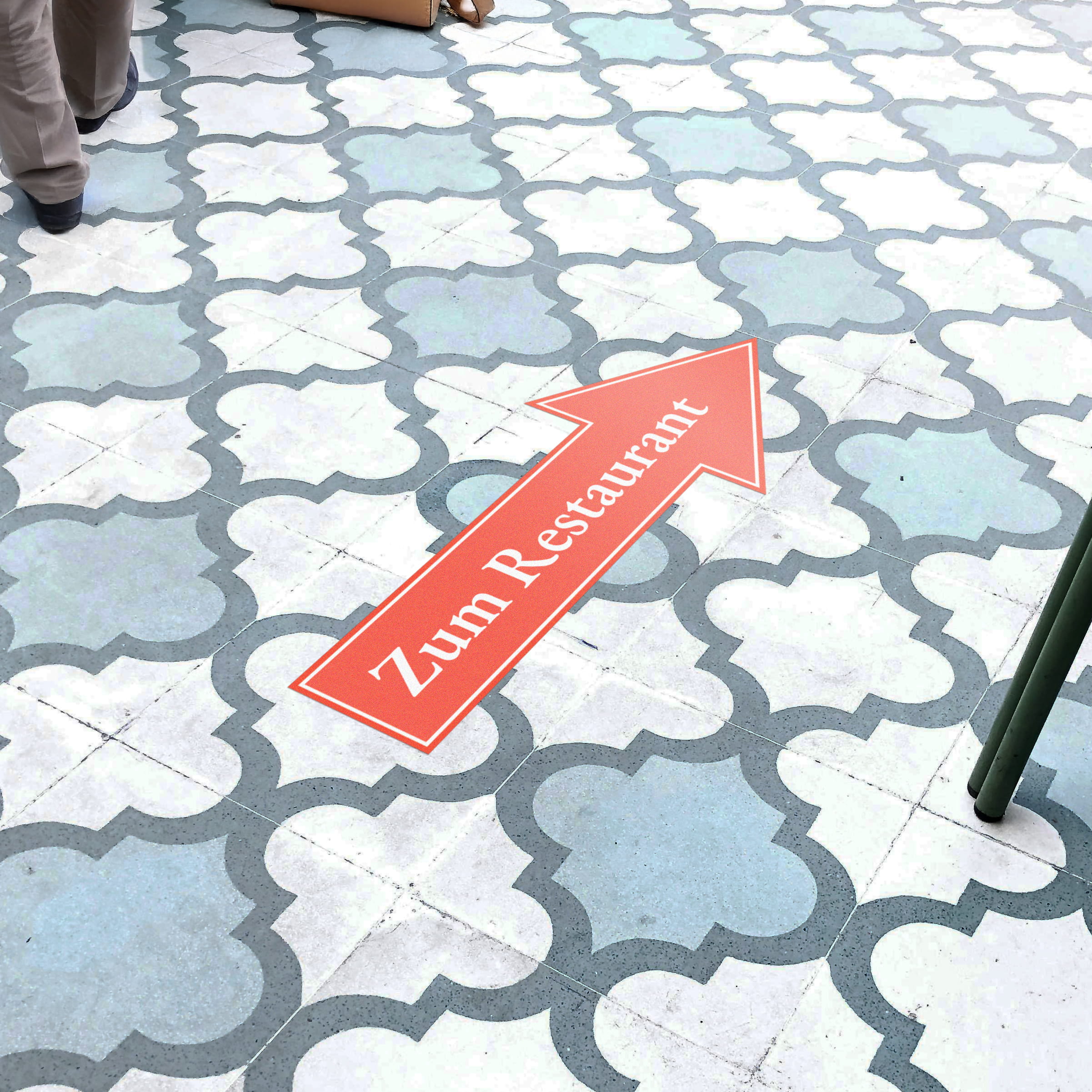 Aufkleber auf dem Boden in Pfeilform mit rotem Hintergrund und weißem Text "Zum Restaurant" auf Fliesen