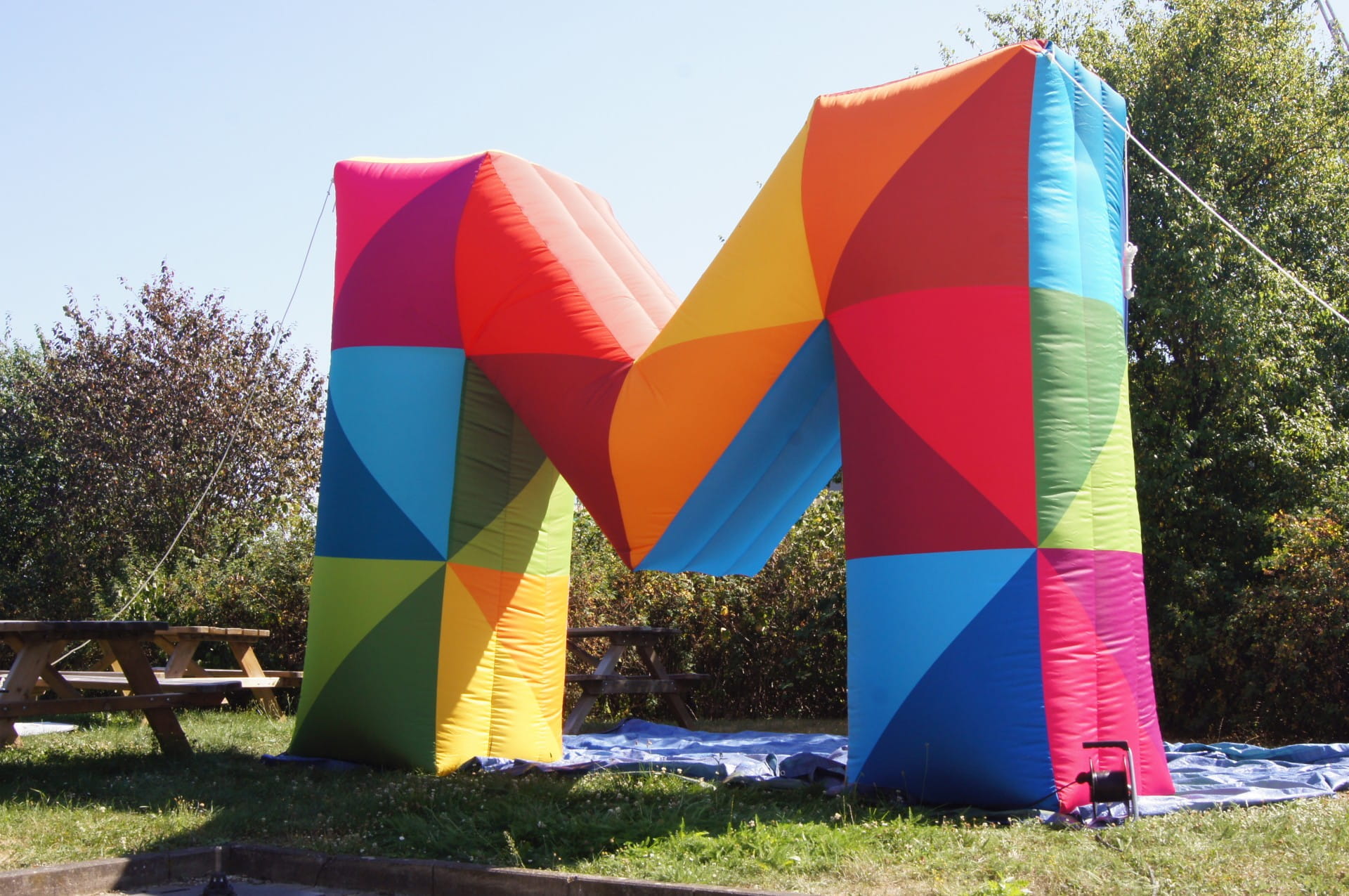 Blow Up Easy Inflate Ad M Buntes, aufblasbares M in verschiedenen Farben, auf einer Wiese mit Bänken im Hintergrund.