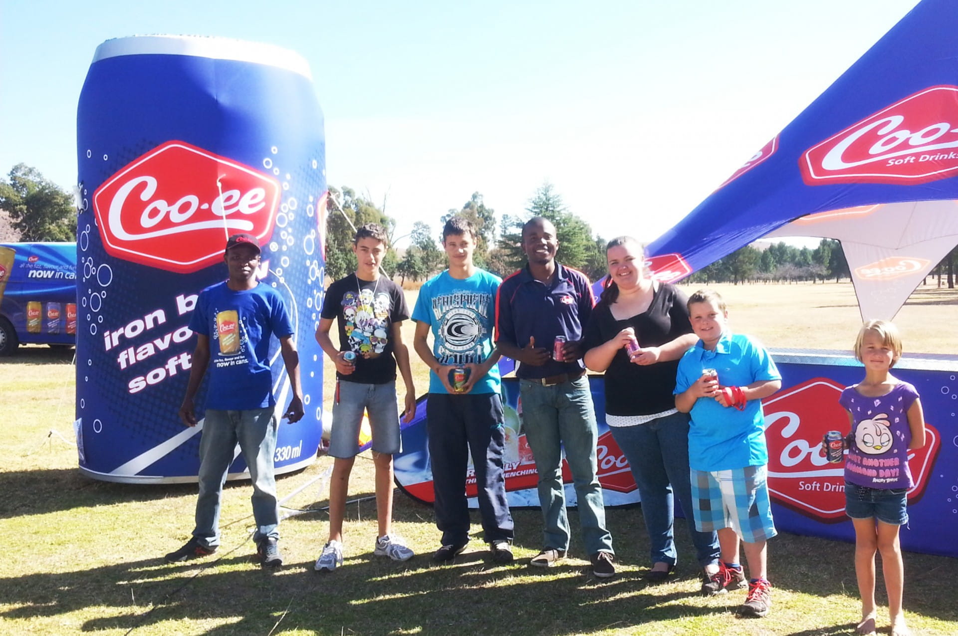 Blow Up Easy Inflate Ad Coo Ee Gruppe von sechs Personen vor einem großen Coo-ee Werbebanner und einem Zelt, im Freien.