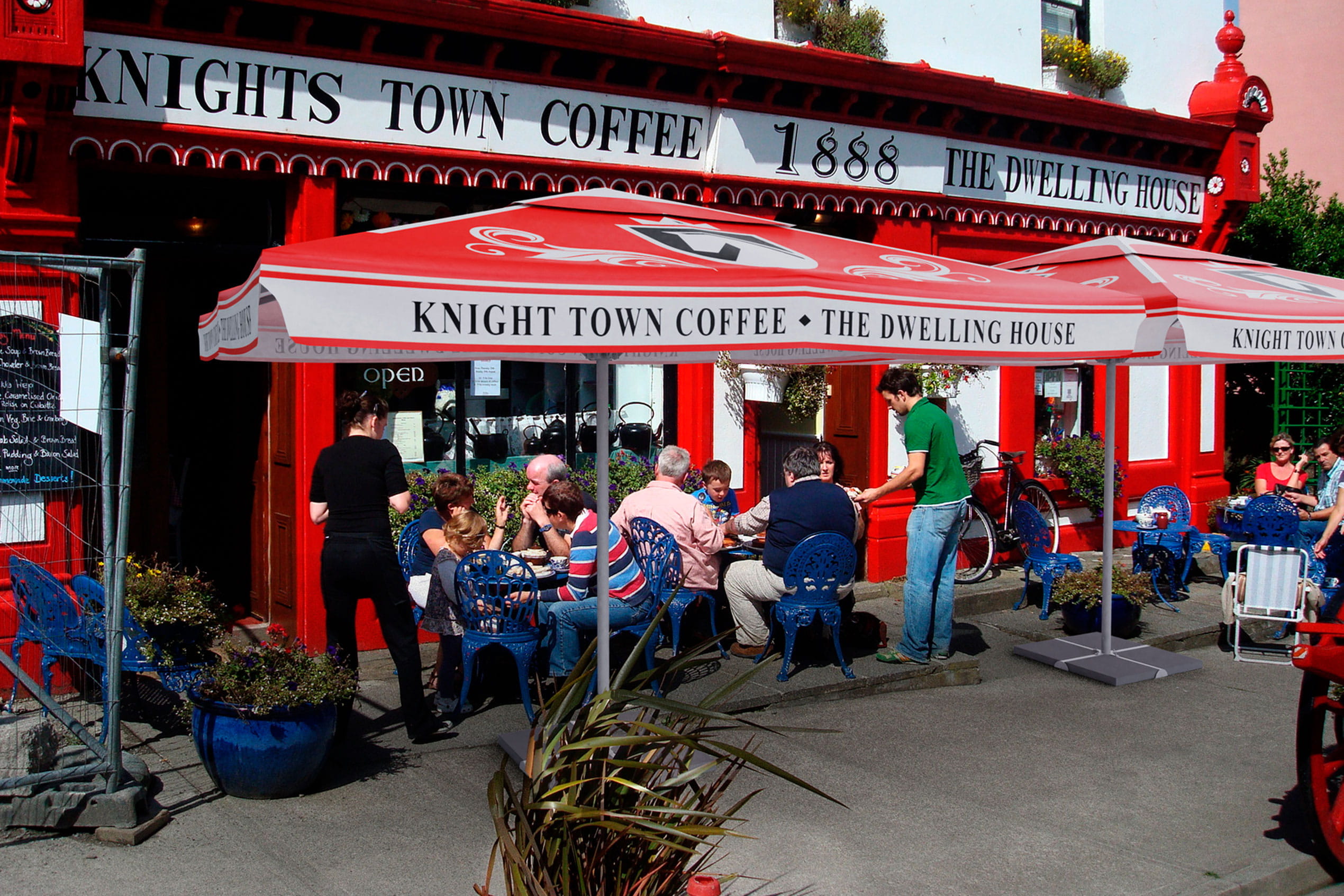 Sonnenschirm Expert Knights Sonnenschirm Expert Knights vor dem Knights Town Coffee, Gäste sitzen an Tischen im Freien.
