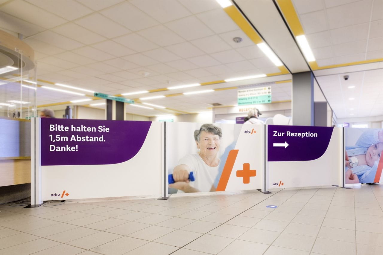 Inhaker Absperrung Absperrung mit dem Hinweis 'Bitte halten Sie 1,5m Abstand. Danke!' und einem Bild einer lächelnden Frau, platziert in einem Empfangsbereich.