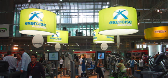 Deckenhänger Cylindrical Beleuchtet Beleuchtete zylindrische Deckenhänger mit dem Schriftzug 'exercise Australia' über einem Fitness-Messebereich.