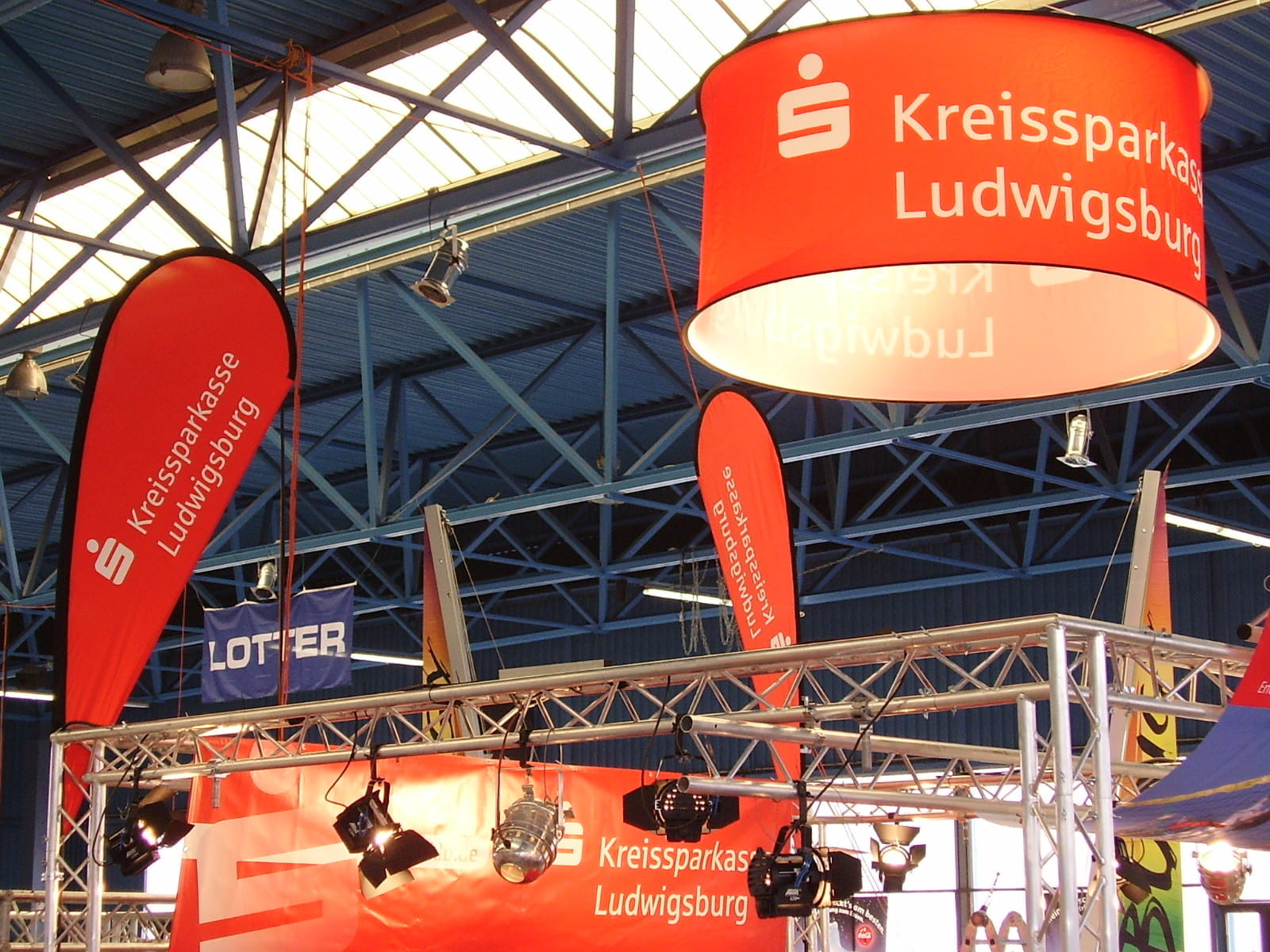 Deckenhänger rund Cylindrical POS Runde Deckenhänger mit dem Toyota-Logo über einem Messestand, Besucher interagieren mit einem Rennwagen.