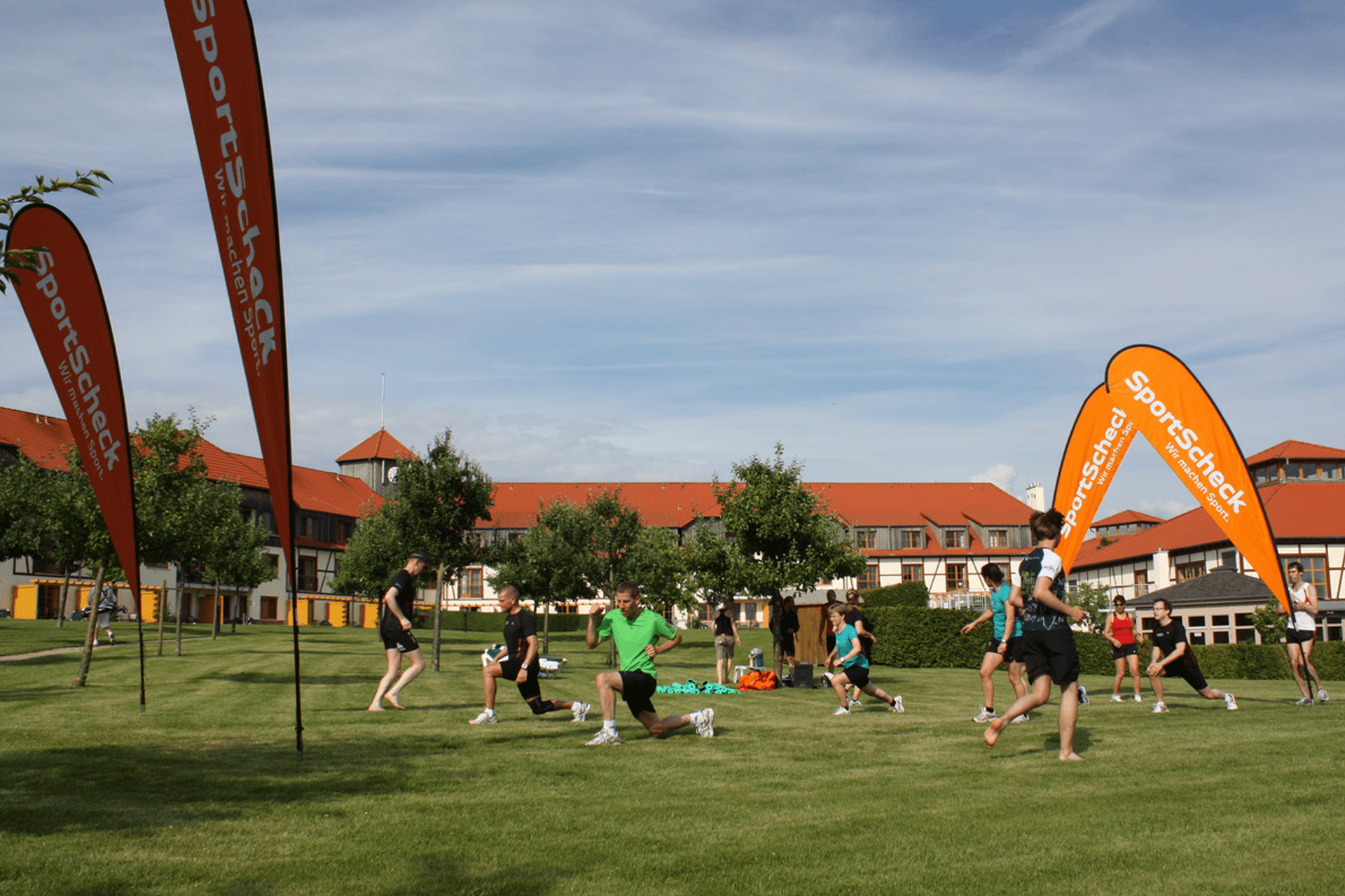 Erwachsene machen Aufwärm- und Dehnübungen auf einer Wiese vor einer Gebäudeanlage mit Beachflags am Rand