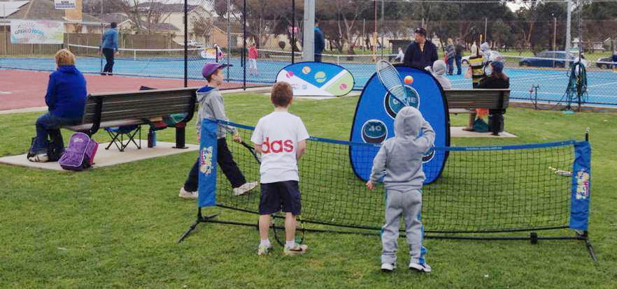 Einsatzbeispiel Quickboard Tennisspiel Zwei Kinder spielen mit einem Tennisball an einem Quickboard Werbebande auf einem Tennisplatz, während Erwachsene im Hintergrund zuschauen.